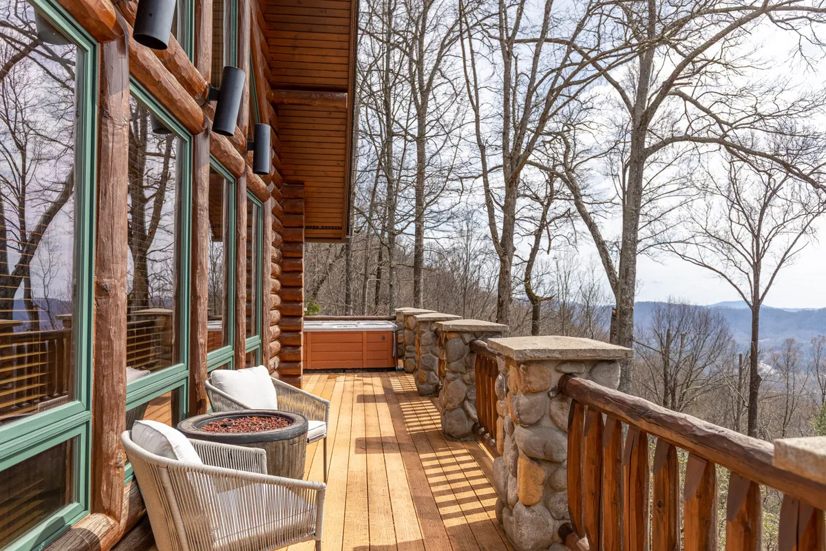 This image captures the log cabin exterior with large windows, a stone railing, a hot tub, and inviting outdoor seating. The cabin is nestled among tall trees, evoking a serene mountain retreat. The scene emphasizes the seamless blend between the comfortable living space and the surrounding natural beauty, inviting guests to step outside and immerse themselves directly in nature.