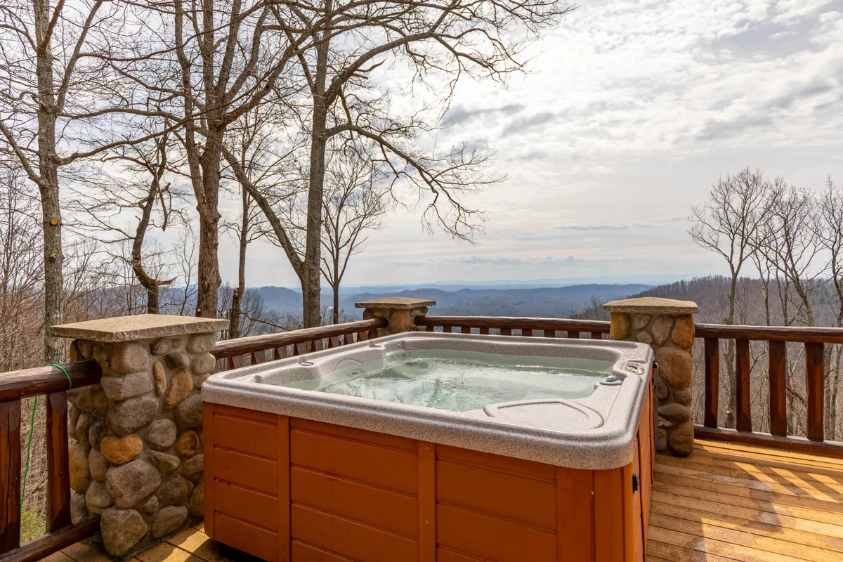 This image showcases a steaming hot tub positioned on a wooden deck with sturdy railings and stone pillars, set against a backdrop of striking mountain views. The hot tub is large enough for a group, and the location offers a secluded, peaceful setting perfect for unwinding and socializing while enjoying the natural beauty of the mountains.