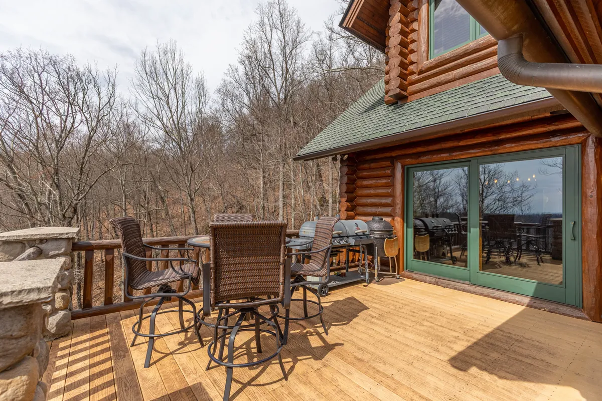 This image showcases the exterior of the log cabin nestled in the woods, featuring an outdoor dining set, grill station, and mature trees surrounding the property. The setting captures the natural beauty and secluded atmosphere of the retreat, with hints of changing foliage and ample outdoor space for seasonal activities.