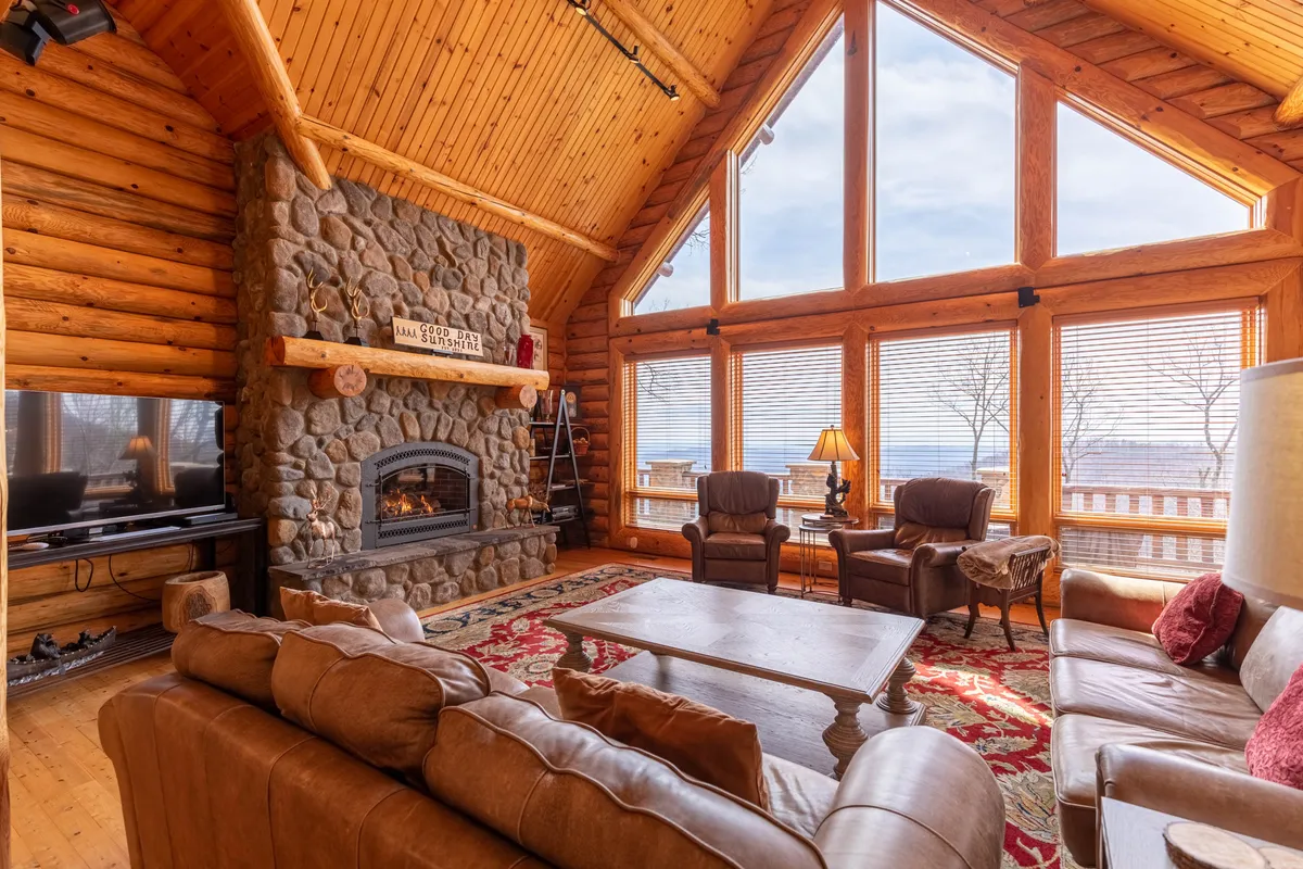 This inviting living room features a large stone fireplace, expansive windows that frame the surrounding wilderness, and a comfortable seating area with a decorative rug. The natural light and panoramic views blend the beauty of the outdoors with the comforts of home.