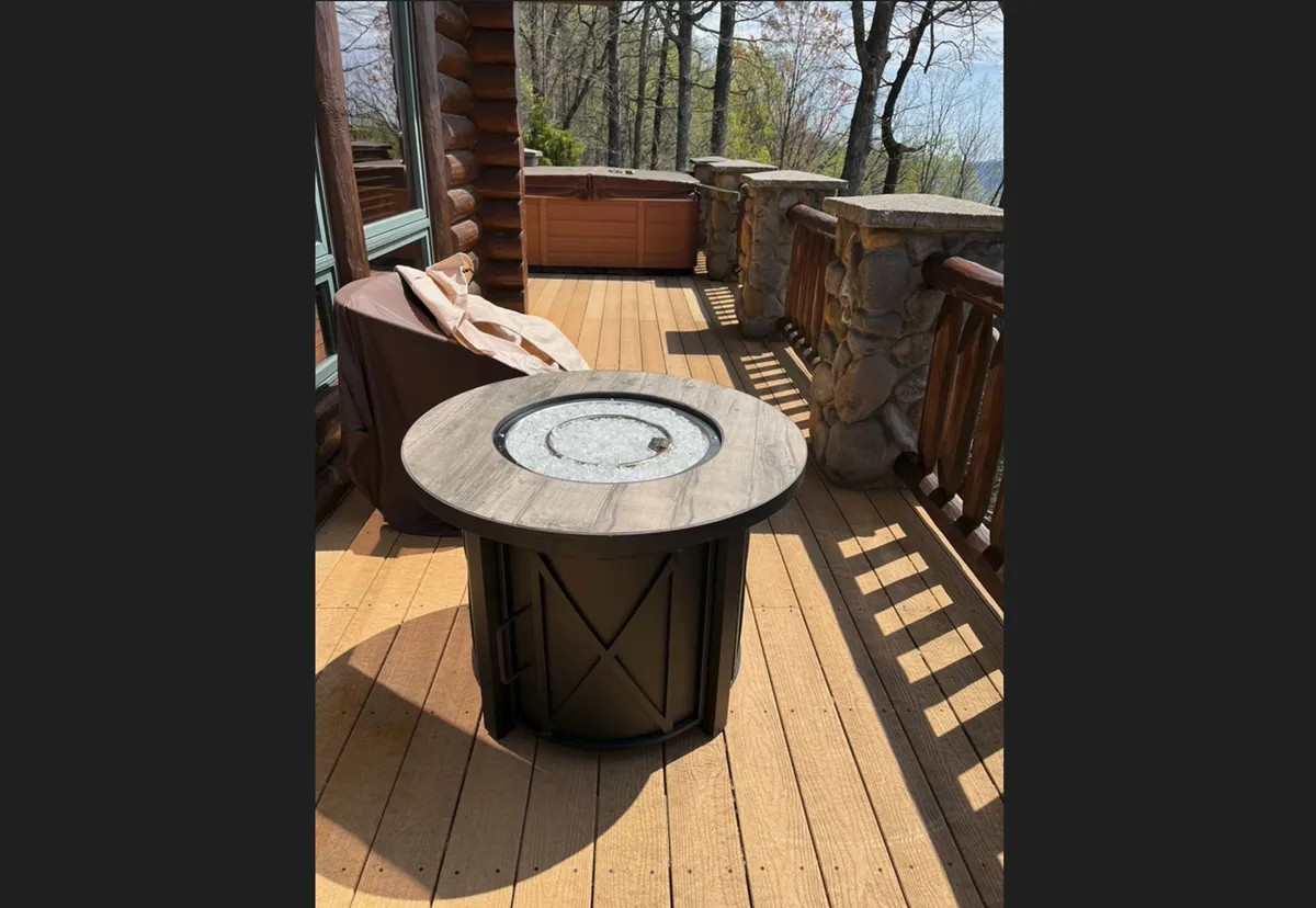 A cozy fire pit table anchors a section of the wooden deck, surrounded by comfortable seating and set against a lush backdrop of trees. The inviting glow of the fire suggests warmth and conviviality, perfect for crisp evenings spent under the stars.