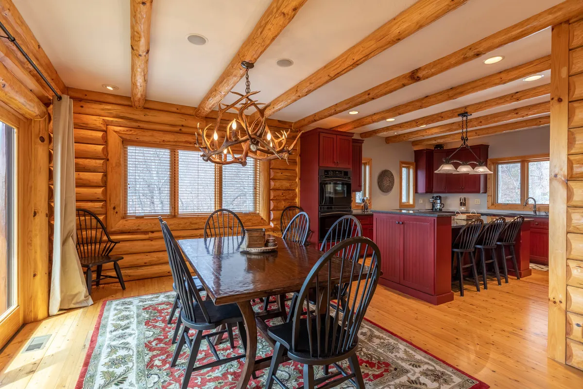 The image features a spacious dining area with a large wooden table, elegant chandelier, and rustic log walls. The kitchen is visible in the background, illustrating the open-concept layout. The setting is perfect for memorable family meals and celebrations.