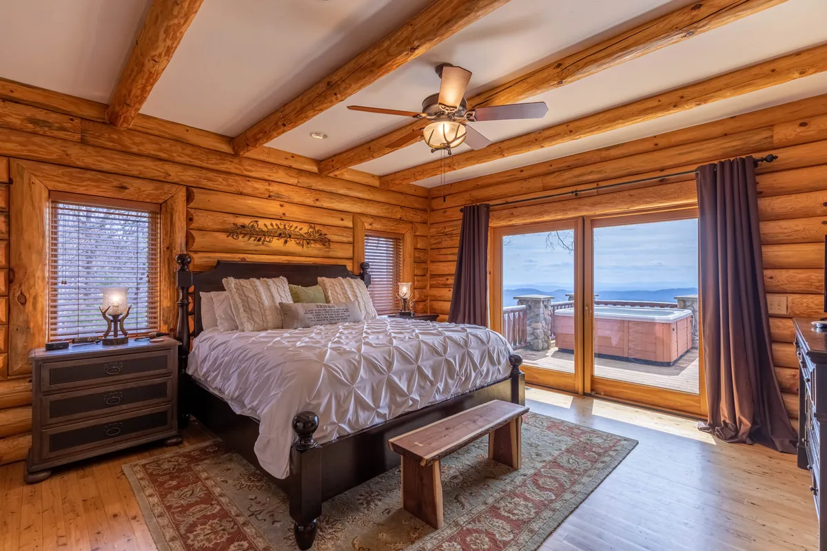 The image presents a cozy bedroom with large windows offering a striking view and a hot tub visible on the deck. The tranquil atmosphere inside is enhanced by the promise of soaking in the hot tub while gazing at the landscape—an inviting prospect, especially in winter after a day of snowy adventures.