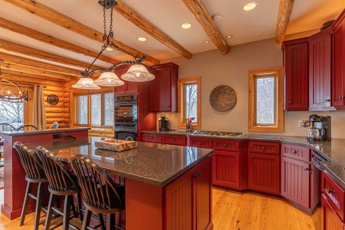 A gourmet kitchen with gleaming granite countertops, warm wooden cabinetry, modern light fixtures, and a set of bar stools for casual gatherings. Large windows let in abundant natural light, enhancing the inviting, functional space ideal for entertaining and meal preparation.