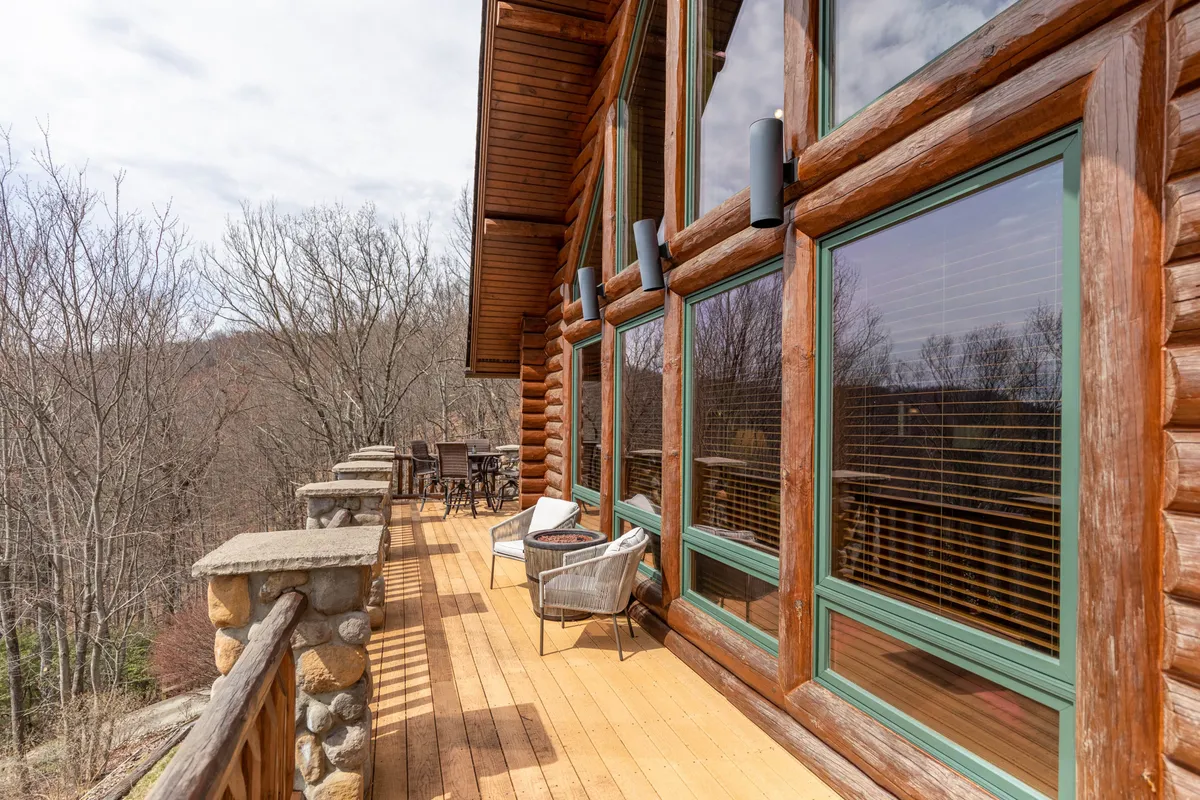 A wide-angle view of the cabin's expansive wooden deck, bordered by a sturdy stone railing, with comfortable outdoor seating arranged to take in the breathtaking multi-layered mountain scenery. The image captures a relaxed atmosphere, with hints of morning mist and a golden sunset sky, suggesting the deck's versatility from sunrise to evening.
