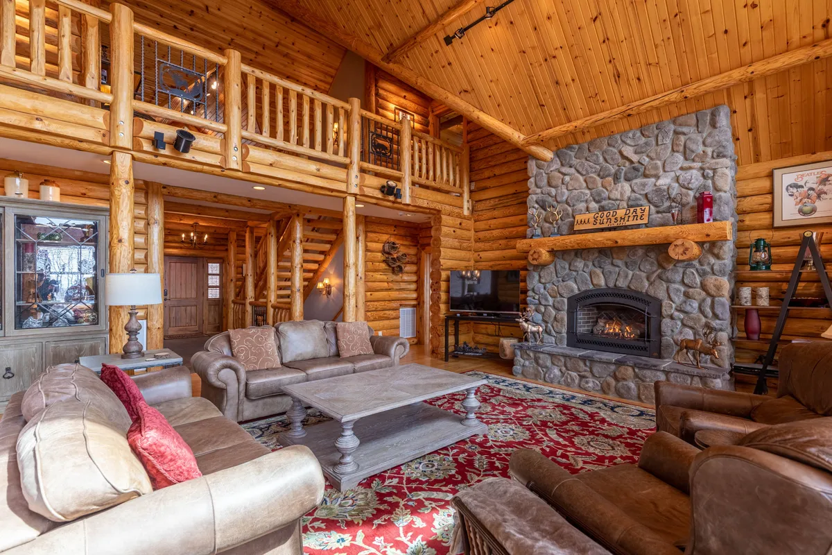 This image showcases a striking stone fireplace framed by wooden beams, with plush, comfortable seating and an ornate rug beneath. The arrangement creates a cozy yet grand centerpiece ideal for family gatherings or evenings by the fire. The high ceilings and natural textures emphasize warmth and luxury.
