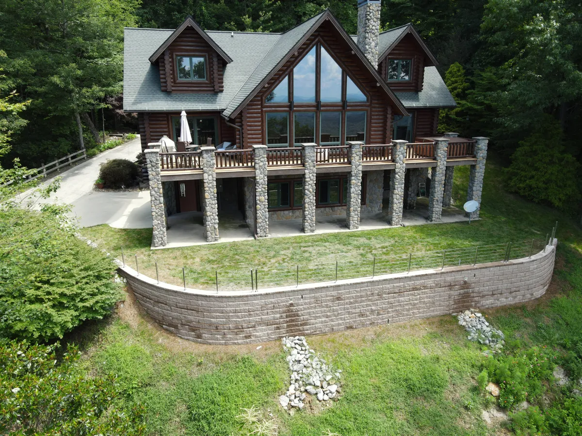 The exterior facade showcases the log cabin's impressive front, featuring large windows, stone columns, and a wooden balcony set against a beautifully landscaped yard. Ample deck space and panoramic views invite outdoor gatherings and moments of quiet reflection at sunrise or sunset.
