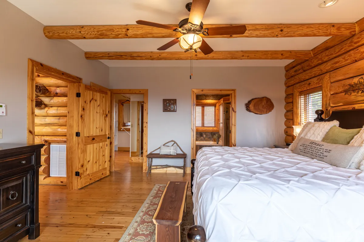 A tranquil bedroom with log walls, decorative pillows on the bed, wooden ceiling beams, and rustic furniture. Sunlight filters through the windows, creating a peaceful retreat perfect for reading or unwinding.