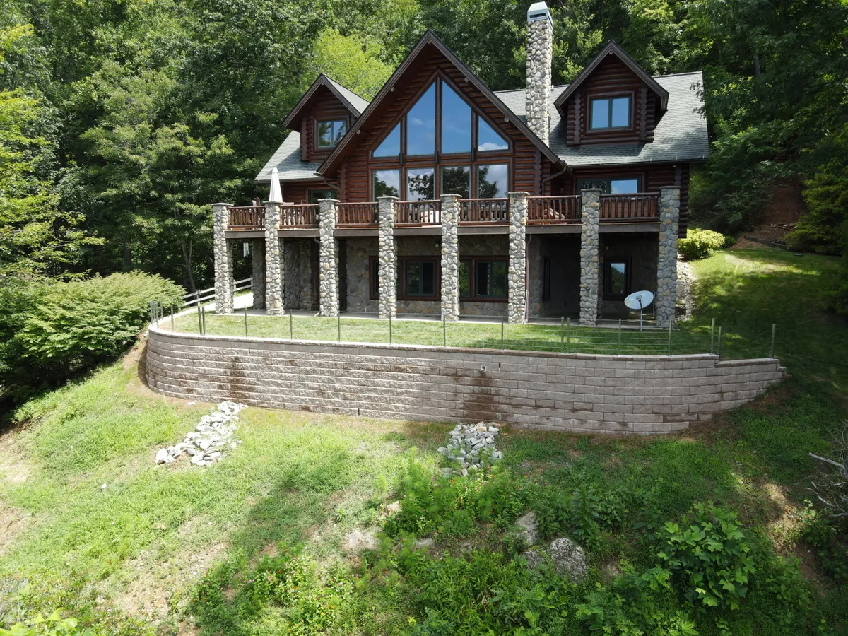 The hero image showcases a beautiful log home nestled against the lush backdrop of Pisgah National Forest. The home features striking large glass windows, sturdy stone pillars, and a spacious wooden balcony. The scene is bathed in warm sunset light, inviting guests to imagine relaxing outdoors, whether sipping morning coffee on the porch, gathering with family on the balcony, or gazing at stars by the fire pit. The expansive acreage surrounding the home hints at the privacy and tranquility awaiting guests.