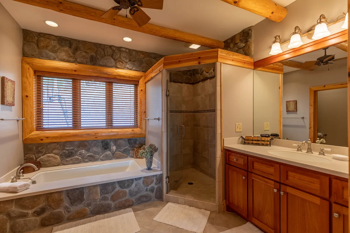A spa-inspired bathroom with a luxurious soaking tub, walk-in shower, vanity with sink, stone wall, and exposed wooden beams. The space feels both tranquil and elegant, ideal for relaxation after a day outdoors.