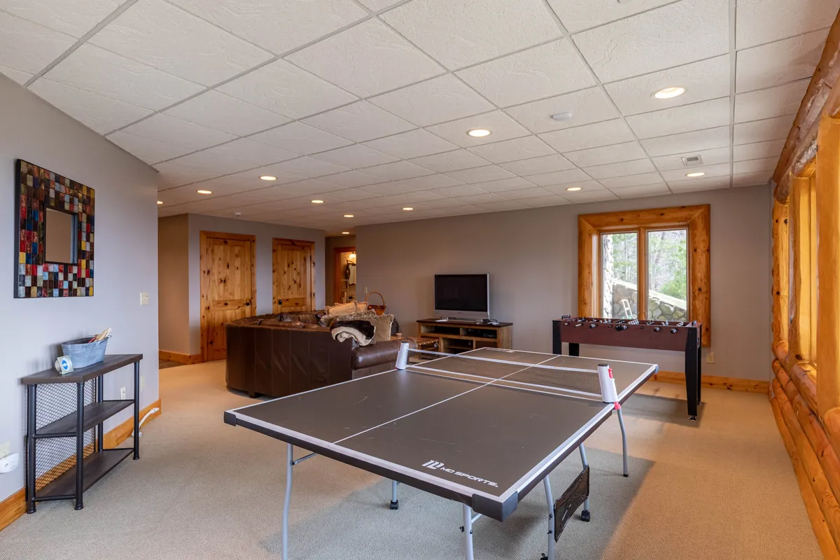 A vibrant lower-level game area featuring a ping pong table and a foosball table, complemented by a large sofa and a wall-mounted television. Natural light filters through a window, and the casual furniture creates a welcoming spot for group play or relaxation.