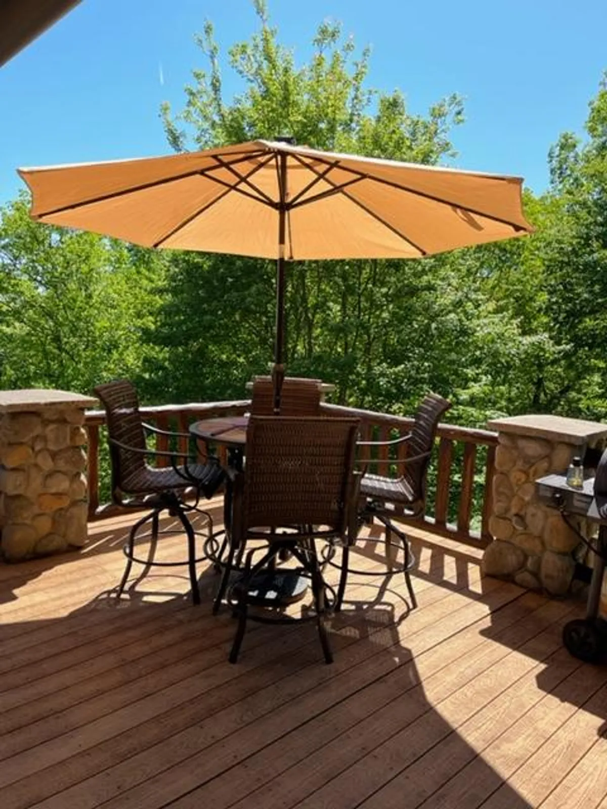 A spacious wooden deck with a round table and chairs under a large umbrella overlooks lush greenery. The inviting setup is perfect for outdoor dining, barbecues, and lounging while taking in mountain views and the shifting light of day.