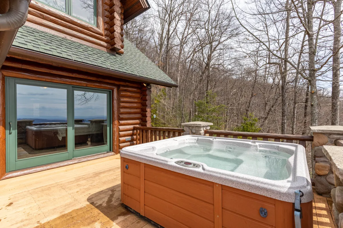 This exterior image captures a luxurious hot tub set on a private deck, framed by the rustic log cabin architecture and surrounded by tranquil forest. The setting evokes relaxation and seclusion, with the hot tub perfectly positioned to take in nature and picturesque sunsets.
