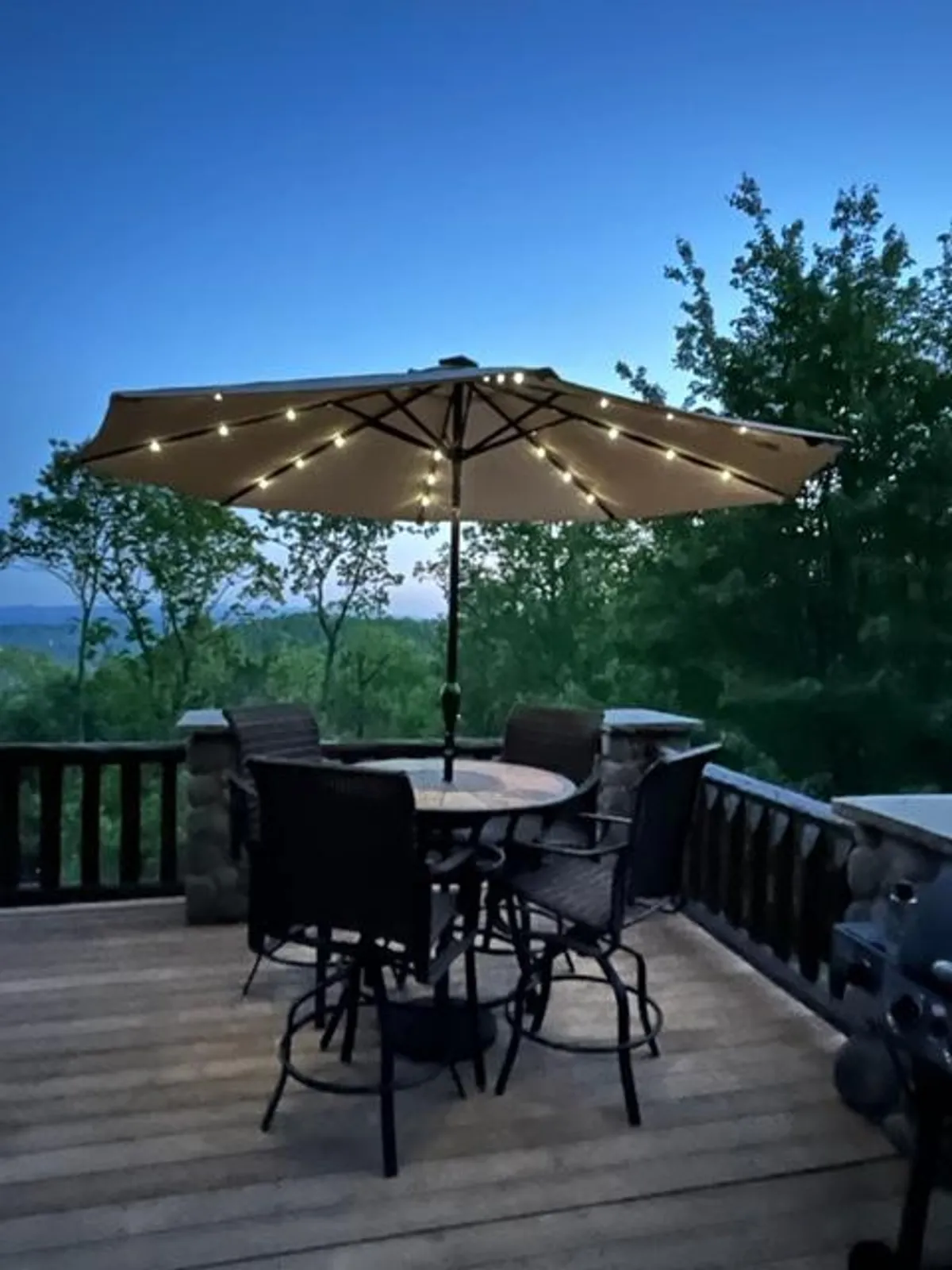 A softly illuminated outdoor dining table sits beneath a glowing umbrella, surrounded by comfortable chairs with a scenic mountain backdrop. The subtle lighting and open setting invite guests to gather for meals, enjoy conversations, or stargaze in a serene, cozy atmosphere.