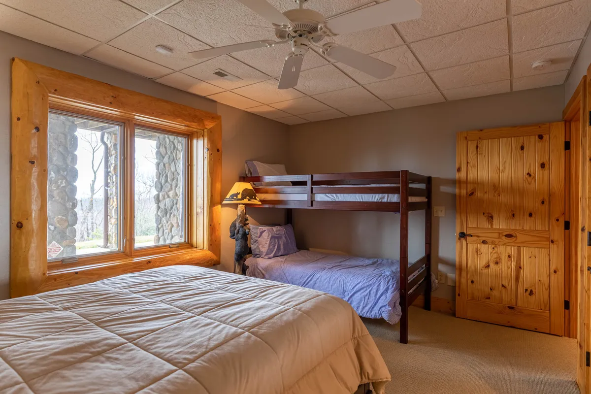 A lower-level bedroom featuring a queen bed and a bunk bed, complemented by a large window that welcomes natural light and wooden accents that add warmth. The setting is family-friendly and inviting, ideal for groups or families with children.