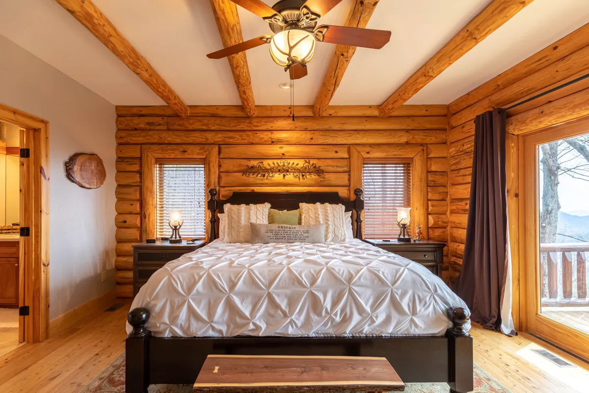 This image features a spacious king-size bed adorned with decorative pillows, wooden beams on the ceiling, and large windows offering mountain views. The nightstands with lamps create a tranquil, cozy ambiance perfect for relaxation.